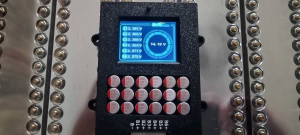 The Heltec 5.5a 6s Balancer and display features an electronics panel with many round, labeled buttons and a small digital screen showing voltage measurements, set against a background of metallic rods and spheres.