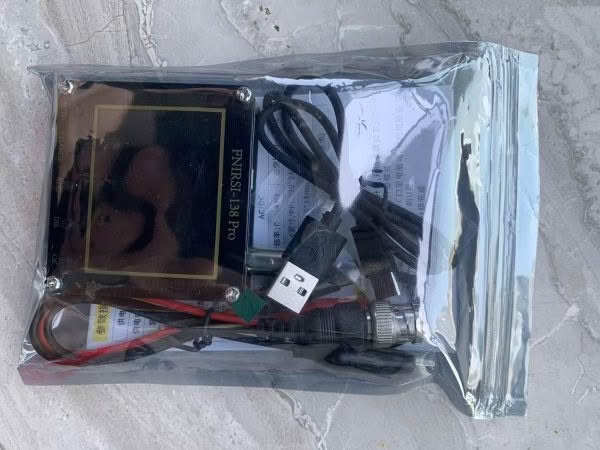 A sealed plastic bag with electronics, including a small black device labeled FNISRI Handheld Oscilloscope, a USB cable, and other wires lies on a textured stone surface.
